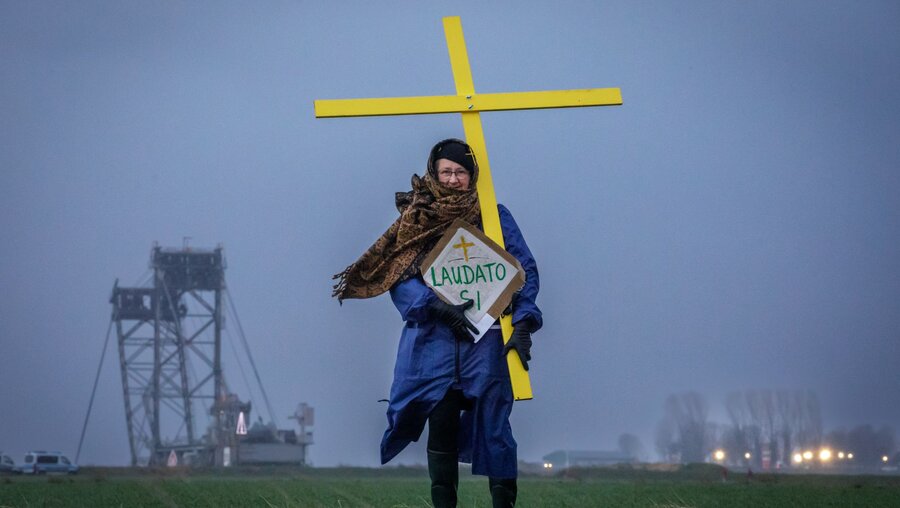 Eine Demonstrantin trägt ein gelbes Kreuz und ein Schild mit der Aufschrift "Laudato Si" am 13. Januar 2023 an der Abbruchkante in Lützerath. / © Gordon Welters (KNA)