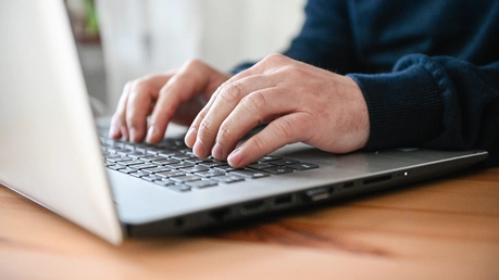 Ein Mann tippt zuhause am Schreibtisch auf der Tastatur von einem Laptop. / © Julia Steinbrecht (KNA)