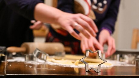 Die Ausstechform für einen Stern steht auf einem Tisch, dahinter ist Teig ausgerollt und Hände stechen Weihnachtsplätzchen aus / © Harald Oppitz (KNA)